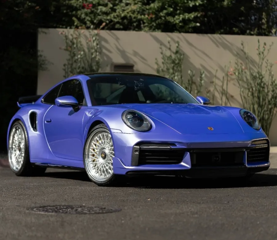 Front three-quarter view of a metallic lavender-blue sports car with polished mesh wheels, parked on asphalt in front of a wall with flowering vines