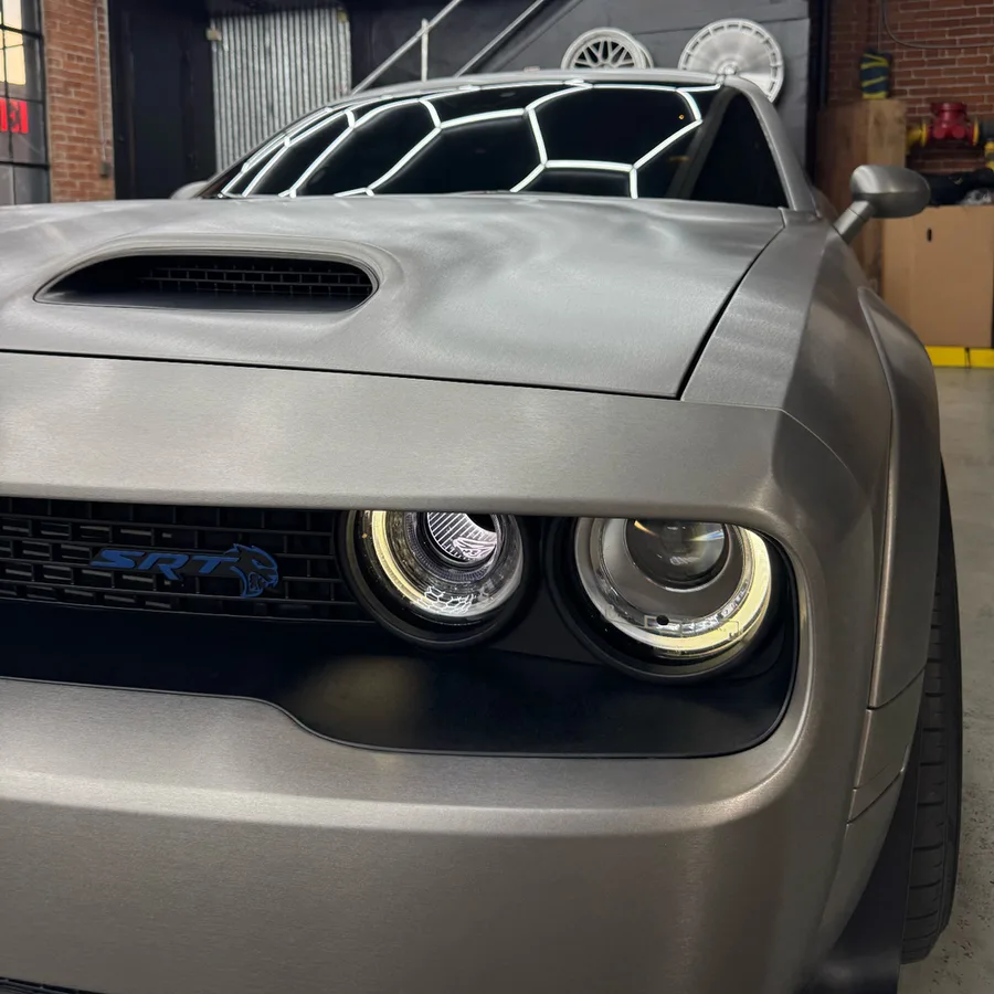 Front end of a muscle car in a brushed metal grey wrap, under the workshop's ceiling lights