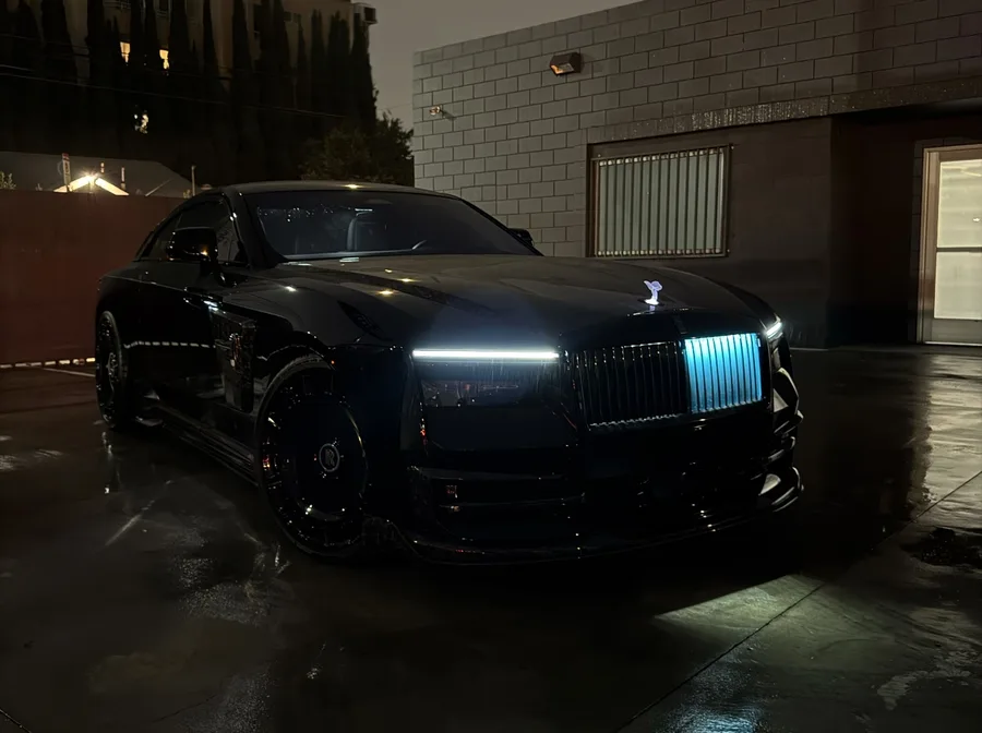 Low front three-quarter view of a gloss black luxury coupe at night, its illuminated grille and headlights glowing on wet pavement